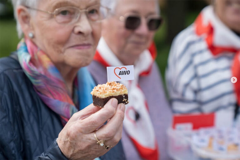 Eine Seniorin hält ein Kuchenstück mit einem kleinen AWO-Fähnchen in der Hand.