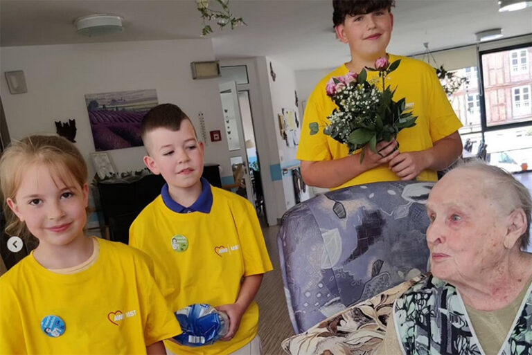 Drei Kinder in gelben Shirts stehen mit einem Blumenstrauß neben einer Seniorin, die im Sessel sitzt.
