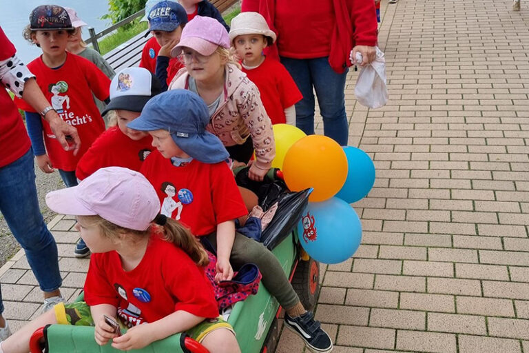 Kita-Kinder mit Käppis und roten Shirts, drei von ihnen sitzen in einem Bollerwagen
