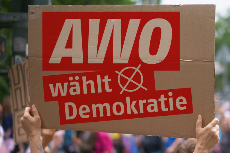 Eine Person, von der nur die Hände zu sehen sind, hält ein Plakat mit der Aufschrift "AWO wählt Demokratie" hoch.