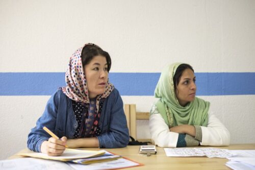 Zwei Frauen mit Kopftüchern sitzen auf Stühlen und blicken nach rechts. Hinter ihnen ist eine weiße Wand mit einem horizontalen hellblauen Streifen