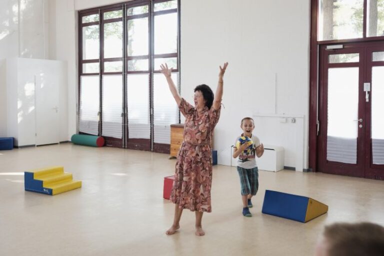 Eine Frau steht in einem Raum und streckt die Hände in die Luft. Hinter Ihr läuft ein kleiner Junge mit Ball.