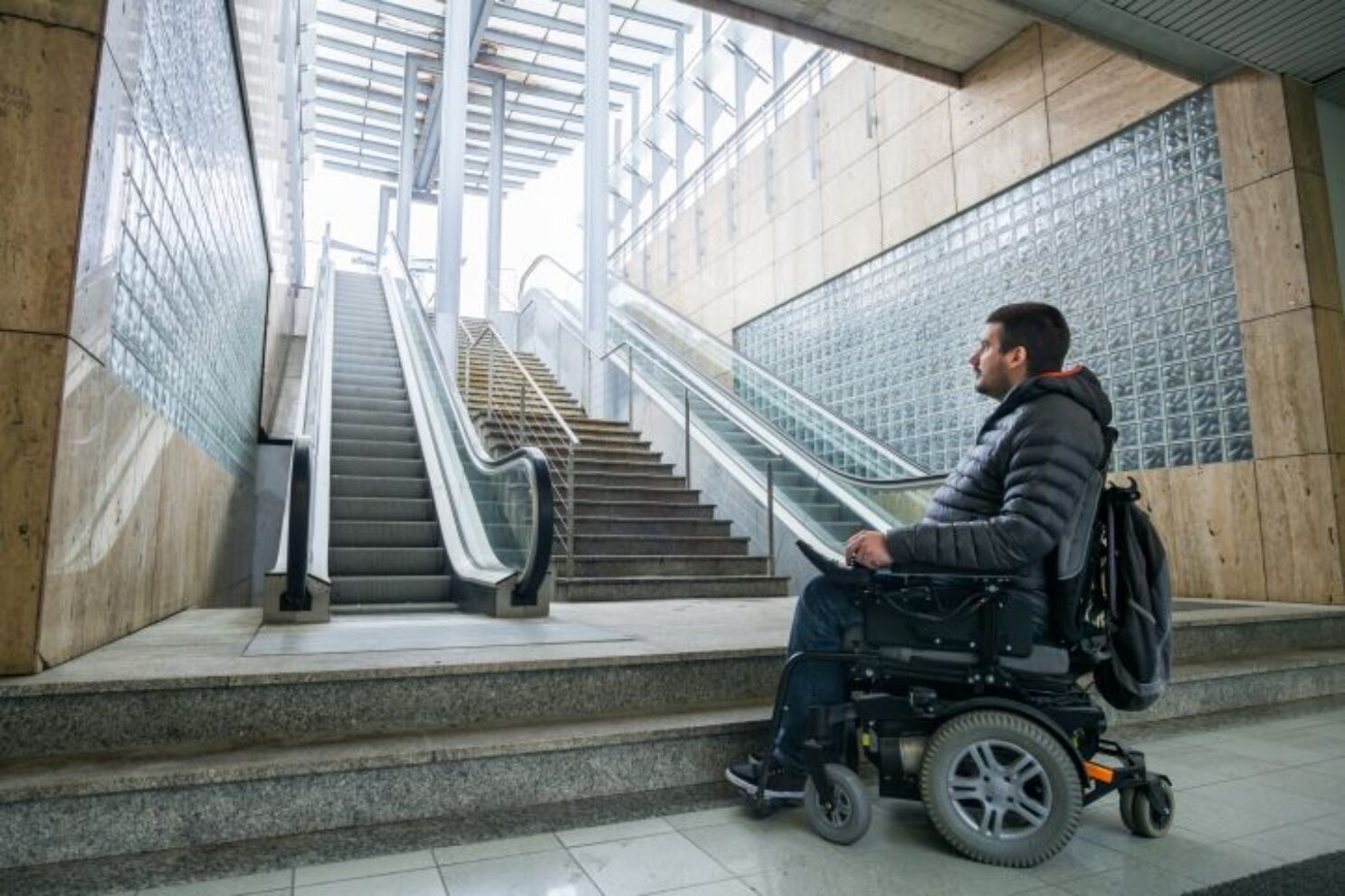 Das Bild zeigt einen Mann der im Rollstuhl sitzt. Er steht am Fuß einer Rolltreppe. Die Rolltreppe ist nicht barrierefrei.