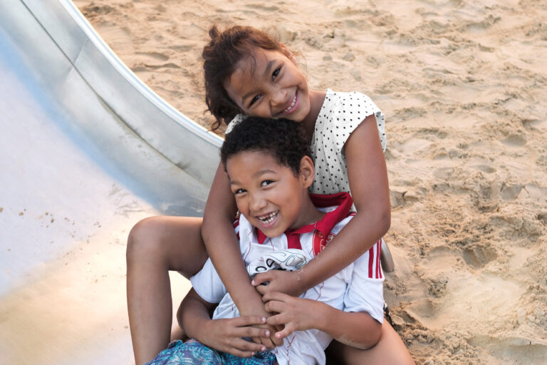 Zwei Kinder (ein Mädche und ein Junge mit Zahnlücke) sitzend fröhlich lachend am unteren Ende einer Rutsche, die in einem Sandkasten steht.