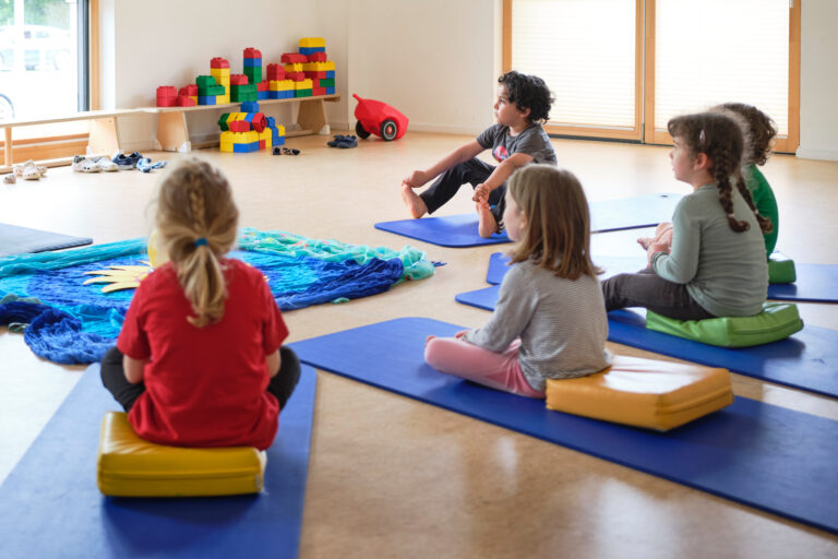 Fünf Kinder sitzen auf Matten in einem Raum, im Hintergrund Lego-Steine und ein Bobbycar-Anhänger