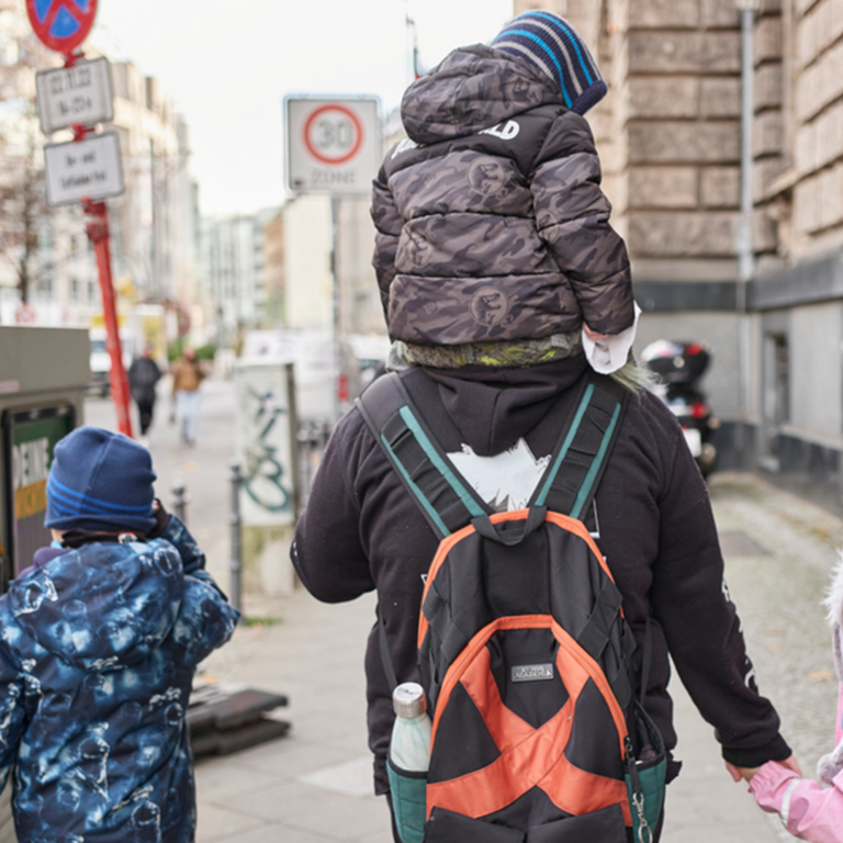 Rückansicht: Erwachsene Person mit einem Kind auf den Schultern und einem Kind an der Hand läuft auf einem Gehweg, daneben ein weiteres Kind.