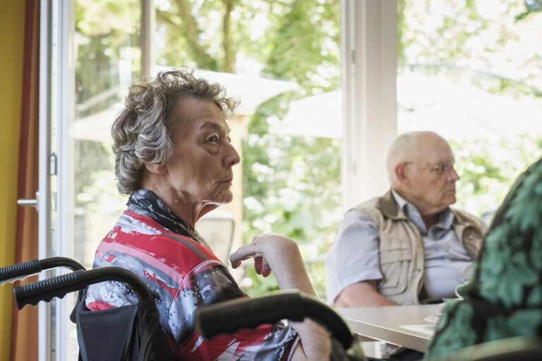 Ältere Personen in einer Pflegeeinrichtung: eine Frau im Rollstuhl und ein Mann sitzen am Tisch.