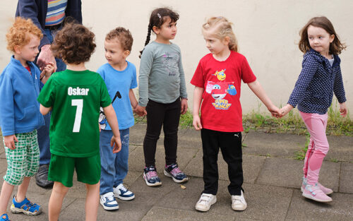 Sechs Kinder - Jungen und Mädchen - halten sich an den Händen und laufen auf einem Gehweg.