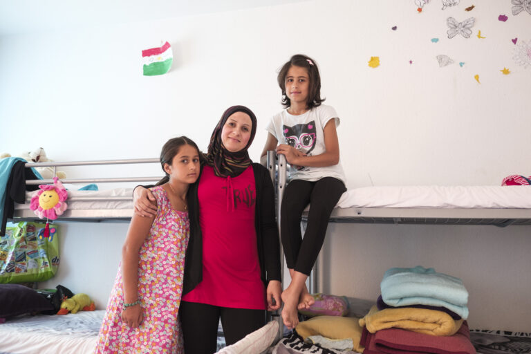 Eine Frau mit Kopftuch steht mit ihren beiden Töchtern vor einem Bett in einer Flüchtlingsunterkunft.