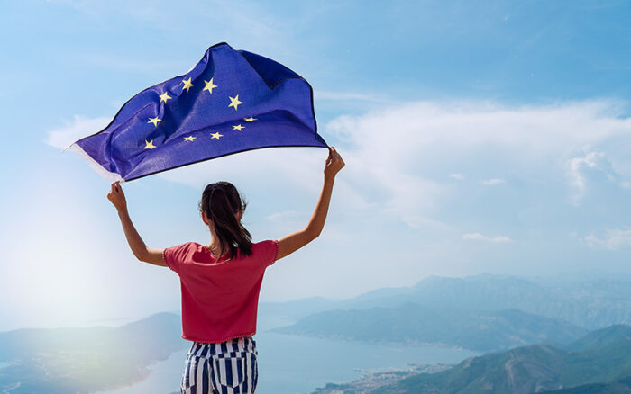 Eine junge Frau steht in der Natur und schwenkt eine Europaflagge über ihrem Kopf.