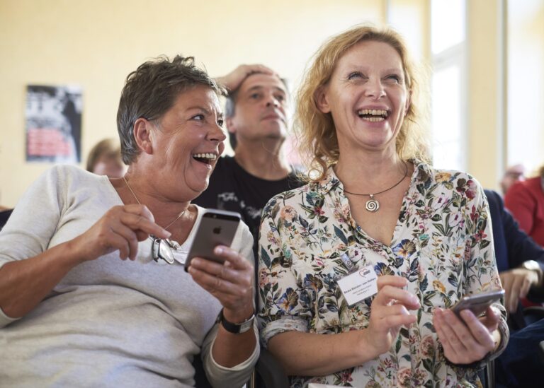 Zwei Frauen sitzen nebeneinander. Sie halten beide ein Smartphone in der Hand und lachen. Im Hintergrund sieht man einen Mann, der sich mit der Hand an den Kopf fasst.