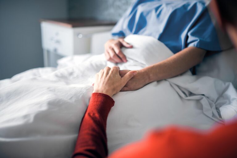 Eine Person, deren Gesicht man nicht sieht, liegt in einem Krankenhausbett. Eine andere Person, die neben dem Bett sitzt, hält ihre Hand.