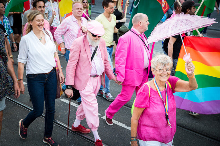 Pride Walk mit Regenbogenflagge, u.a. eine Seniorin mit rosa Schirm und Regenbogenschlüsselband um den Hals und zwei Senioren, die sich an der Hand halten