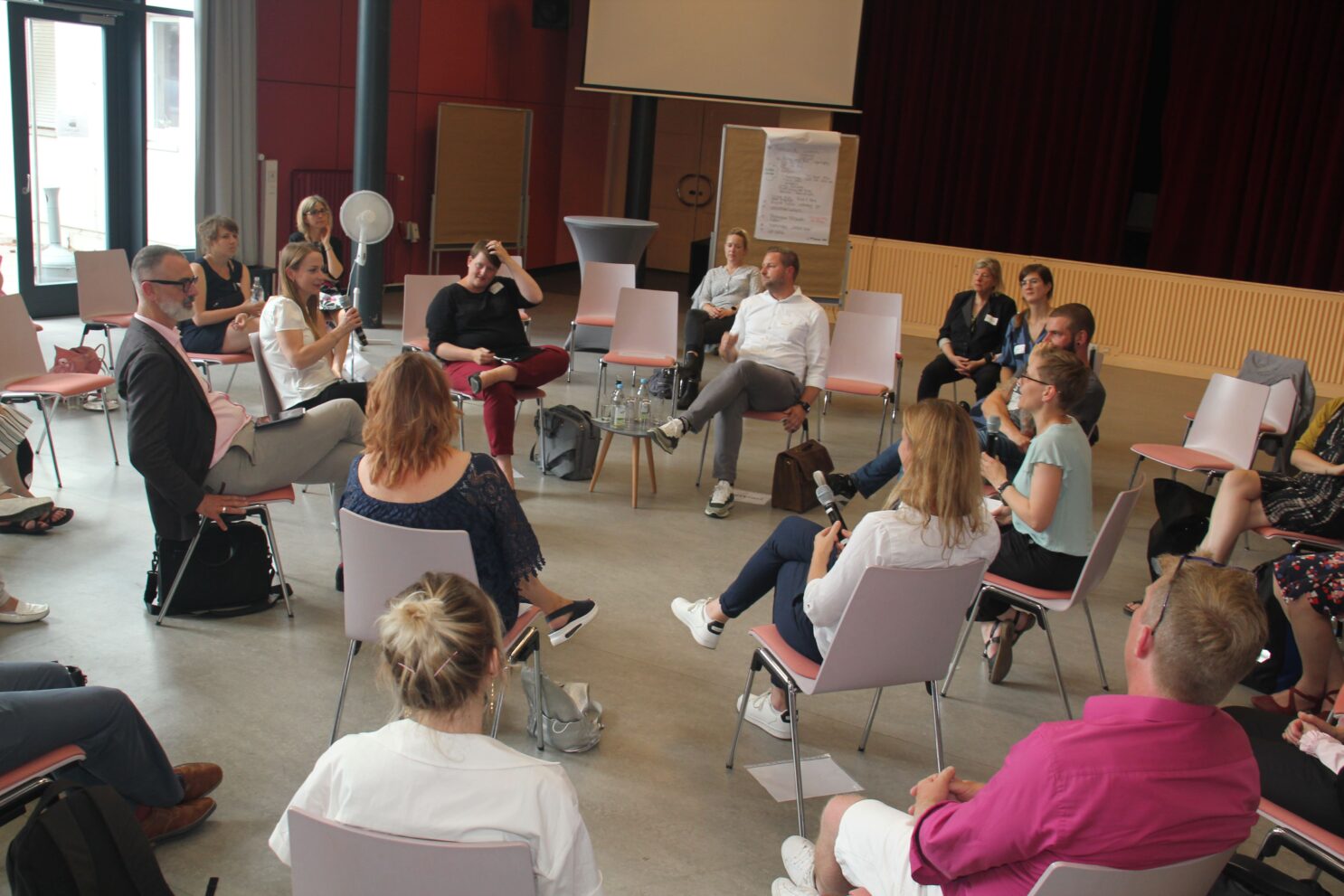 In einem großen Saal sitzen viele unterschiedliche Leute in einem großen Stuhlkreis zusammen. Eine Frau hält ein Mikrofon und spricht in dieses hinein.