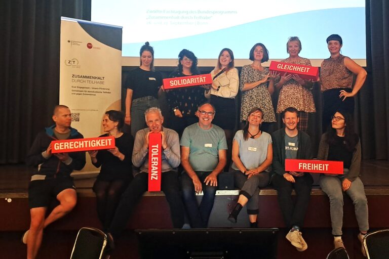Auf einer Bühne stehen und sitzen 15 Personen. Sie halten rote Blöcke in den Händen auf denen Solidarität, Gleichheit, Gerechtigkeit, Toleranz und Freiheit stehen.