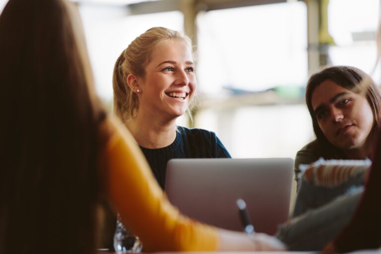 Eine junge lächelnde Frau sitzt an einem Tisch mit anderen Personen. Vor ihr steht ein geöffneter Laptop.