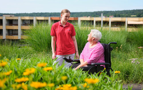AWO-Mitarbeiterin im roten Shirt mit AWO-Logo neben Seniorin im Rollstuhl