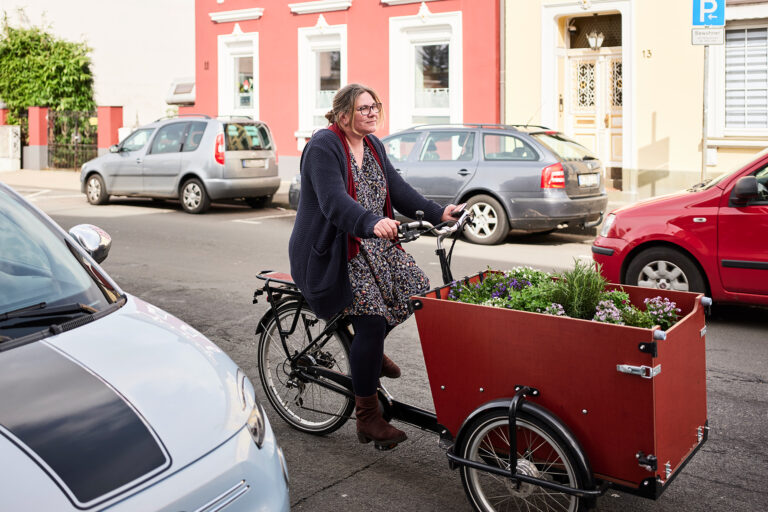 Frau auf einem mit Pflanzen beladenen Lastenrad auf einer Straße zwischen parkenden Autos