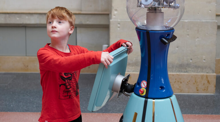 Ein Junge im roten Pullover steht in einem Museum am Display eines blauen Roboters.