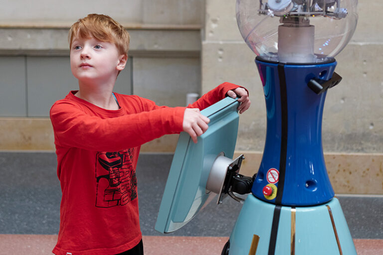 Ein Junge im roten Pullover steht in einem Museum am Display eines blauen Roboters.