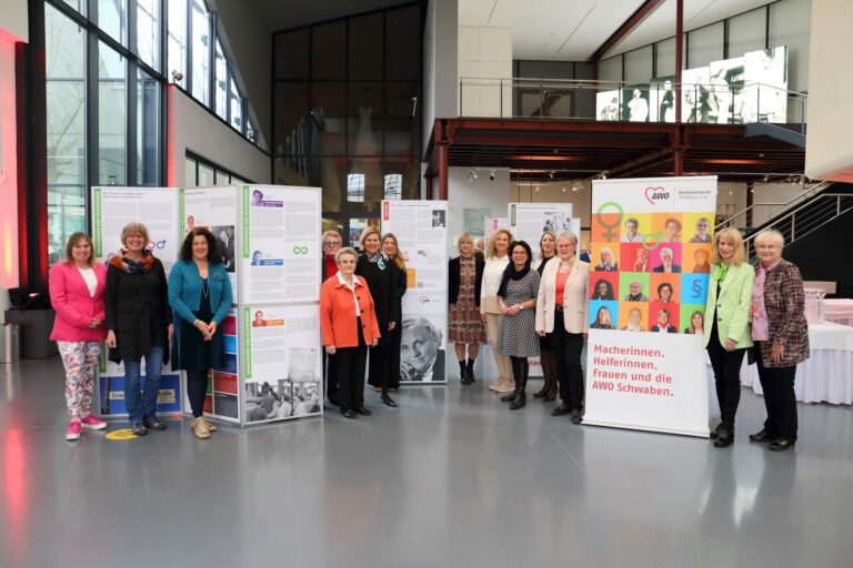 Bei der Ausstellung „Macherinnen. Helferinnen. Frauen und die AWO Schwaben“ posieren einige Frauen für ein Gruppen-Foto . Zwischen ihnen stehen AWO Rollups.