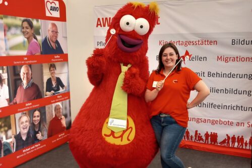 Vor einem AWO-Stand posiert eine Frau in rotem AWO-Shirt neben einem großen, roten Maskottchen mit gelber Krawatte. Sie halten beide kleine AWO Windmühlen.