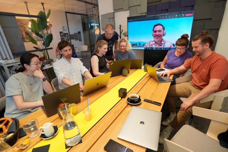 In einem Meeting-Raum sitzen Personen mit ihren Laptops. auf einem Bildschirm im Hintergrund ist eine Person per Video-Konferenz hinzugeschaltet.