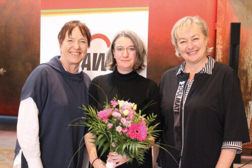 Drei Frauen lächeln in die Kamera. Eine von ihnen hält einen Blumenstrauß in der Hand.
