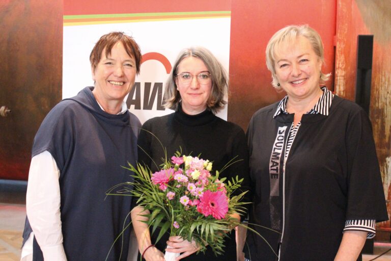 Drei Frauen lächeln in die Kamera. Eine von ihnen hält einen Blumenstrauß in der Hand.