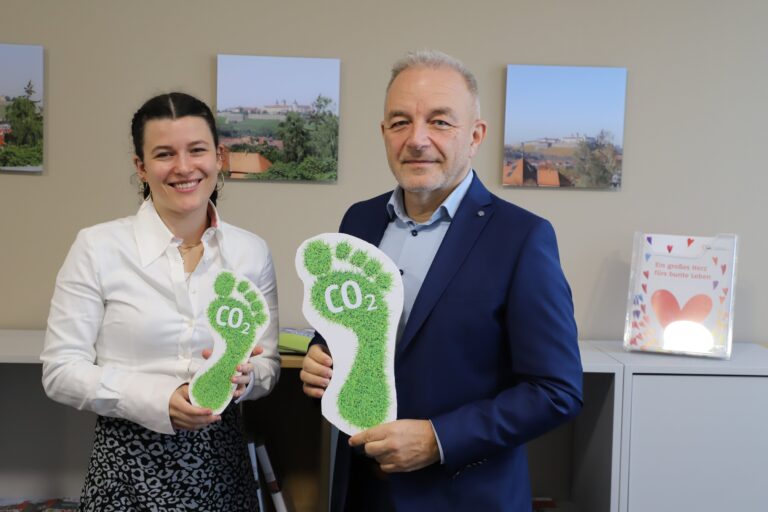 Eine junge Frau und ein Mann halten zwei Schilder mit einem aus grünem Gras bestehenden Fußabdruck in dem CO2 steht.