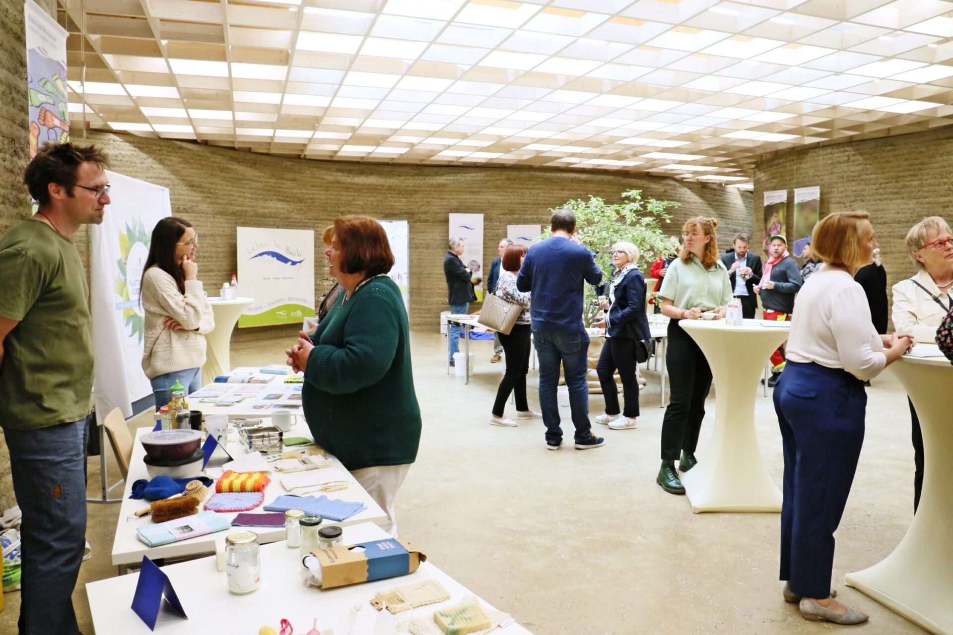 In einem Raum stehen Menschen an Stehtischen und unterhalten sich. Zudem gibt es Tische auf denen diverse Sachen liegen. unter anderem Auch Flyer.