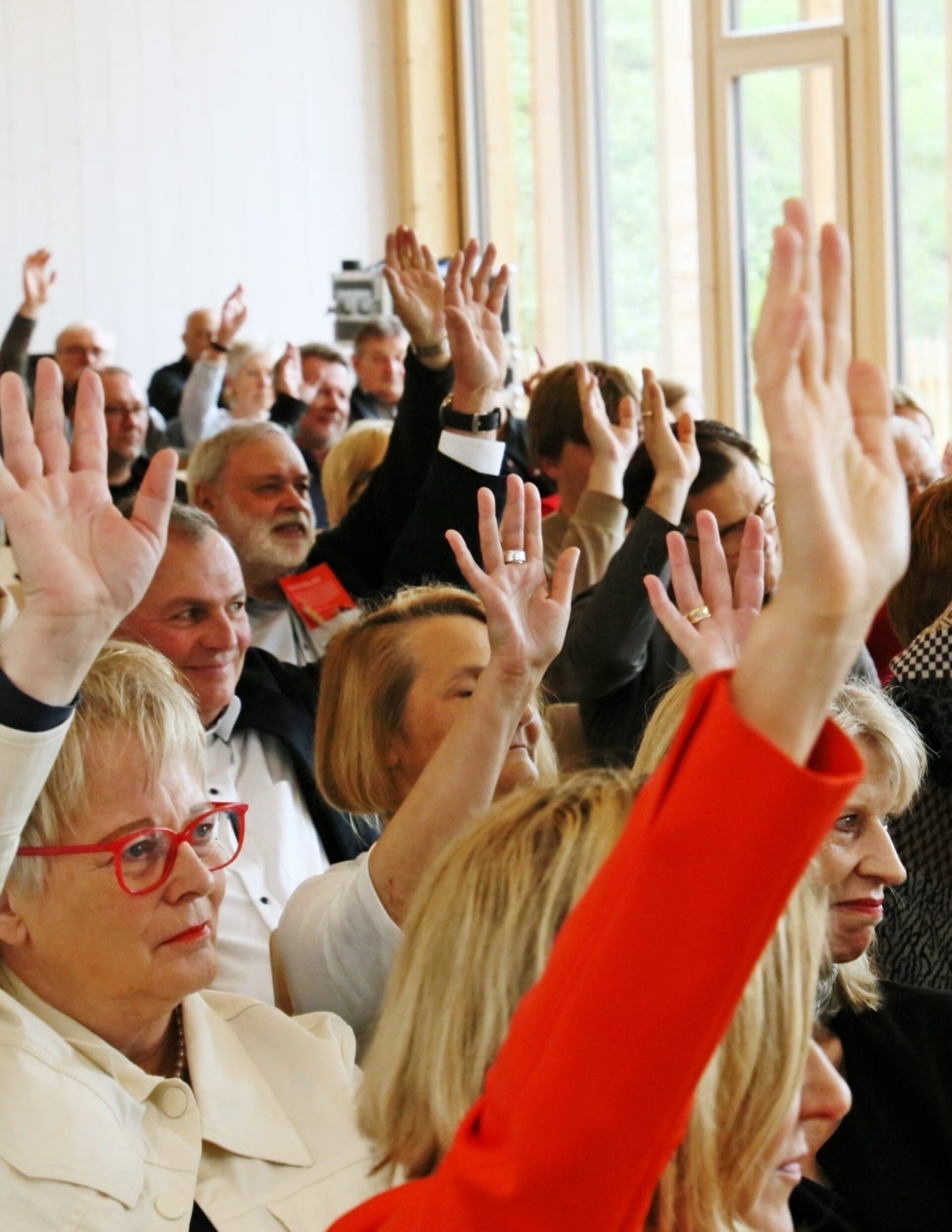 Eine Gruppe von Menschen sitzen in einem Raum. Viele von ihnen heben ihre Hand zur Abstimmung.