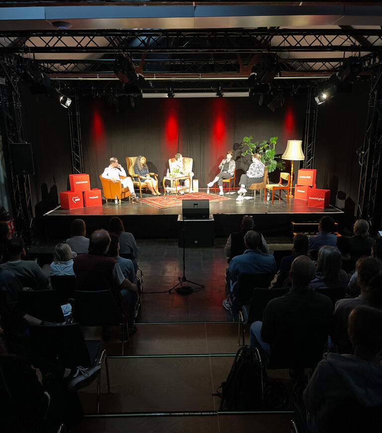 Podiumsdiskussion: Ein kleiner Saal mit einer Bühne. Auf der Bühne sitzen fünf Personen in Sesseln.