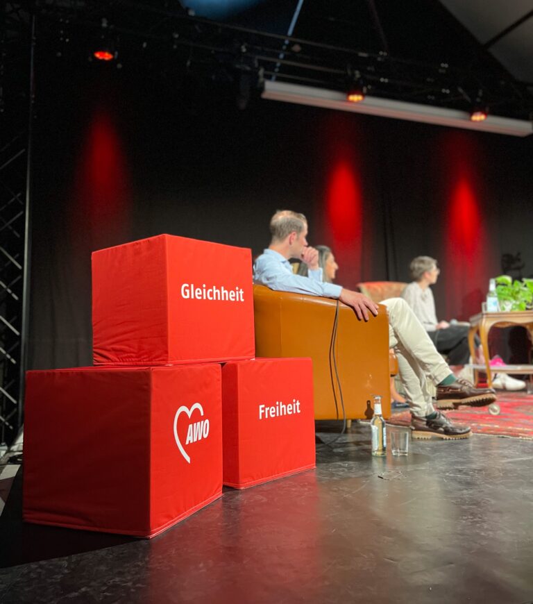 Podiumsdiskussion: Auf einer Bühne sitzen mehrere Personen. Im Vordergrund stehen drei große rote Würfel, die mit Gleichheit, Freiheit und dem AWO-Logo beschriftet sind.