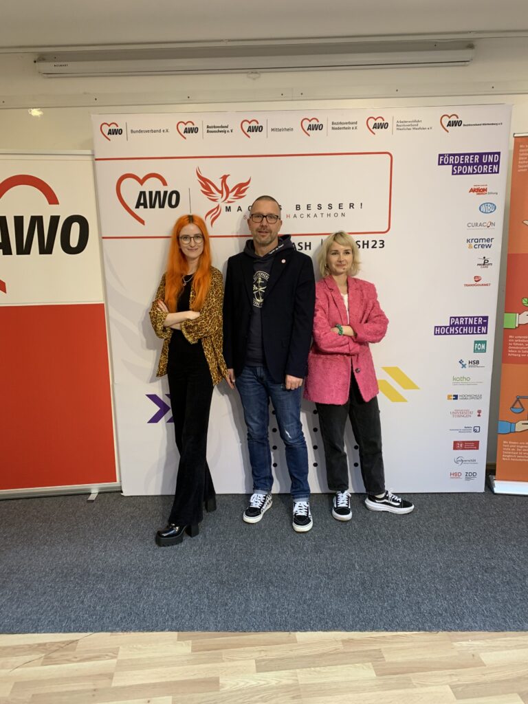 Drei junge Personen in Alltagskleidung stehen vor Rollups der AWO.