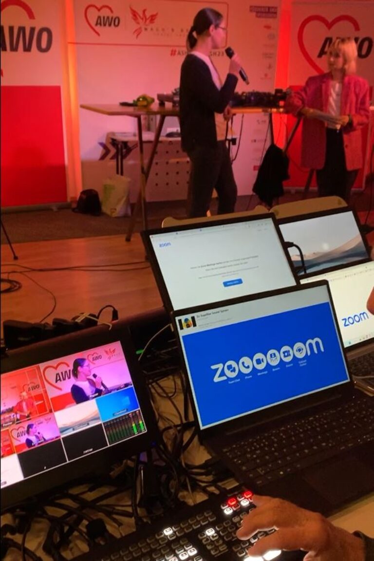 Auf einem Tisch stehen Laptops auf denen verschiedene Programme zu sehen sind. Im Hintergrund führt eine Frau ein Interview mit einer anderen jungen Frau.