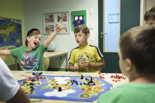 Fünf Jungs spielen in einem Klassenzimmer an einem Tisch ein Brettspiel. Auf dem Tisch liegen viele Figuren und Spielkarten.