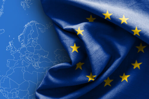 Nahaufnahme der Europaflagge, die mit gelben Sternen auf blauem Hintergrund. Im Hintergrund befindet sich eine stilisierte Landkarte von Europa.