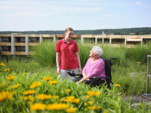 Eine jüngere Frau mit rotem AWO-Shirt und eine ältere Frau im Rollstuhl stehen bzw. sitzen auf einer grünen Wiese.