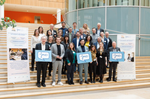 Eine Gruppe von ca. 25 Personen steht auf einer Treppe. Sie lächeln in die Kamera und halten blaue Schilder mit dem JMD Logo.