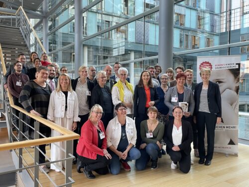 Eine Gruppe von ca. 30 Personen steht vor und auf einer Treppe. Sie lächeln in die Kamera. Neben ihnen steht ein Rollups auf dem steht "Bündnis Kinder Grundsicherung".