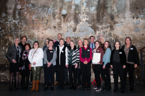 Eine Gruppe von ca. 20 Personen steht vor einer mit Graffiti besprühten Wand. Alle lächeln direkt in die Kamera.