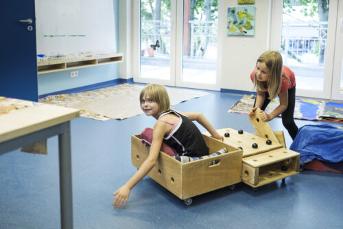 Zwei Kinder spielen in einem Zimmer mit Holzkisten auf Rädern.