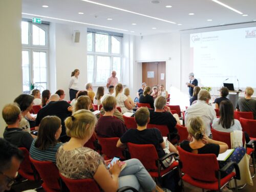 Ein Konferenzraum, in dem viele Menschen in roten Stühlen sitzen und einem Vortrag zuhören. Auf einer Leinwand im Hintergrund ist das Logo der Arbeiterwohlfahrt (AWO) zu sehen.