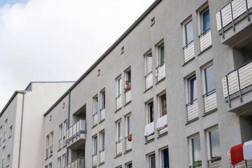 Aus einer Häuserfront mit vielen Fenstern und einigen Balkonen schaut ein Mann aus einem Fenster.