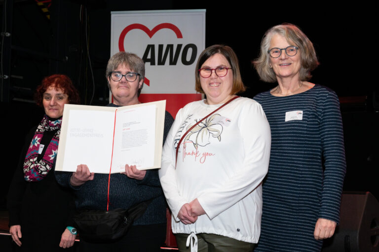 Vier Frauen stehen vor einer Bühne auf dem ein AWO-Rollup steht. Eine der Frauen hält eine weiße Urkunde.