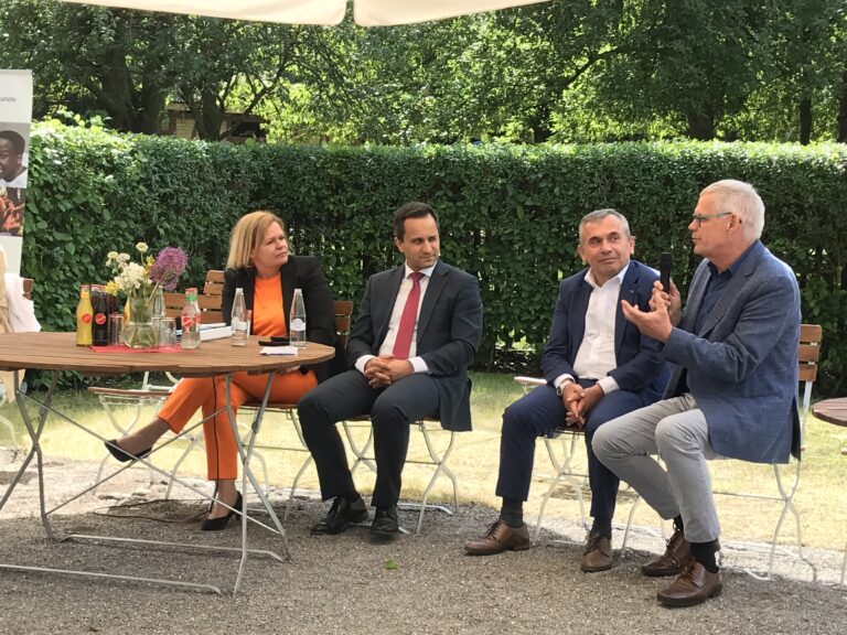 Vier Personen sitzen draußen auf Gartenstühlen. Ein Mann hält ein Mikrofon in der Hand und redet.