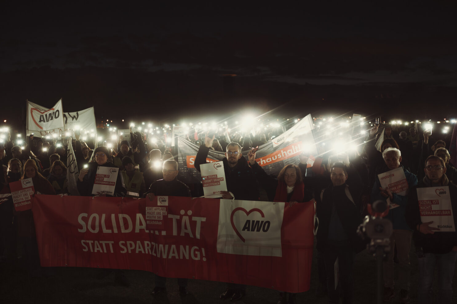 Viele Menschen stehen draußen im Dunkeln, halten Lichter in den Händen sowie AWO-Flaggen und Banner. Auf dem größten Banner steht "Solidarität statt Sparzwang!"