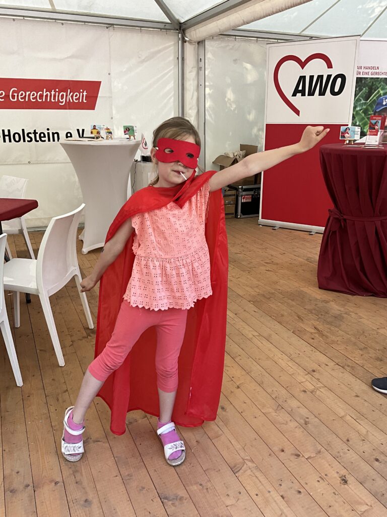 Ein junges Mädchen posiert in einem Zelt mit AWO-Logos, gekleidet als Superheldin mit rotem Umhang und Maske, den linken Arm streckt sie in die Luft.