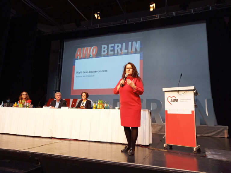 Eine Frau hält auf einer Bühne mit Mikrofon eine Rede vor einem Publikum, während sie neben einem Rednerpult steht. Im Hintergrund ist eine Leinwand mit der Aufschrift 'AWO Berlin' und 'Wahl des Landesvorsitzes' zu sehen. Auf der Bühne sitzen mehrere Personen an einem Tisch.